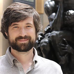 Matt Moser with brown beard and button-up shirt smiles in front of a black statue