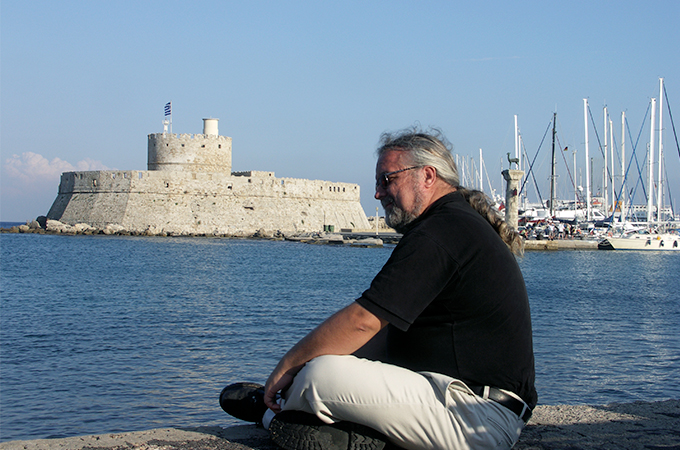 Kelly Devries sits on a stone wall overlooking a harbor, with a stone fort in the background