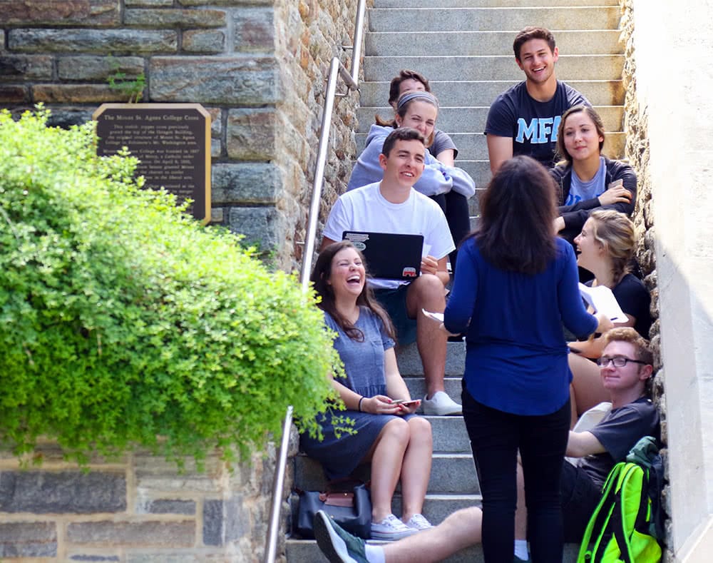A professor teaches a small class outdoors, with the students sitting on concrete stairs in front of her