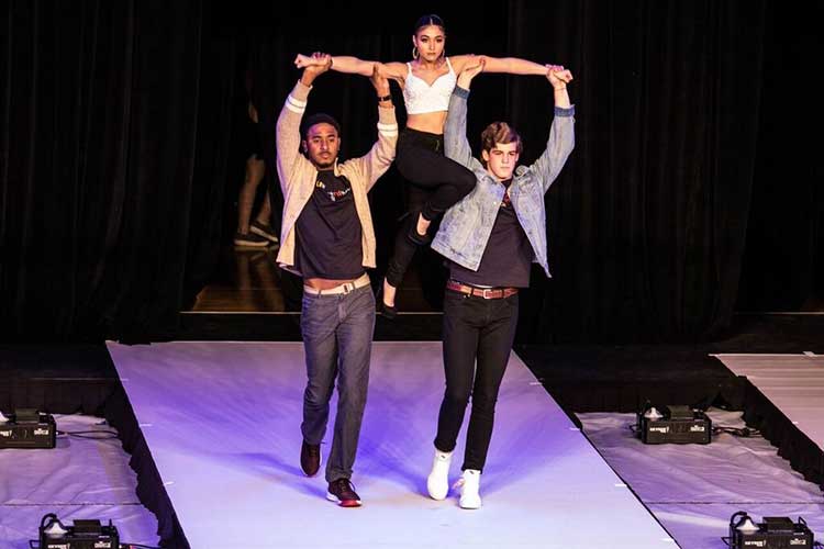 A female model being lifted in the air by her arms by two male models on the runway