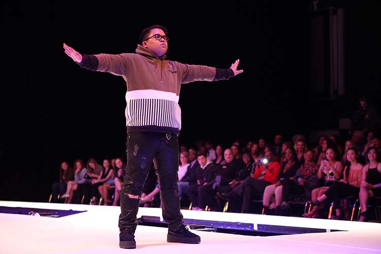 A male model standing alone at the end of the runway in a color-block outfit with arms stretched out to the sides