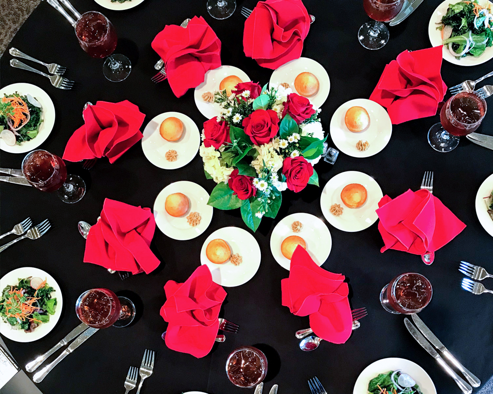 Aerial view of a table set with bread, salad, and iced tea featuring a red rose centerpiece and red folded napkins