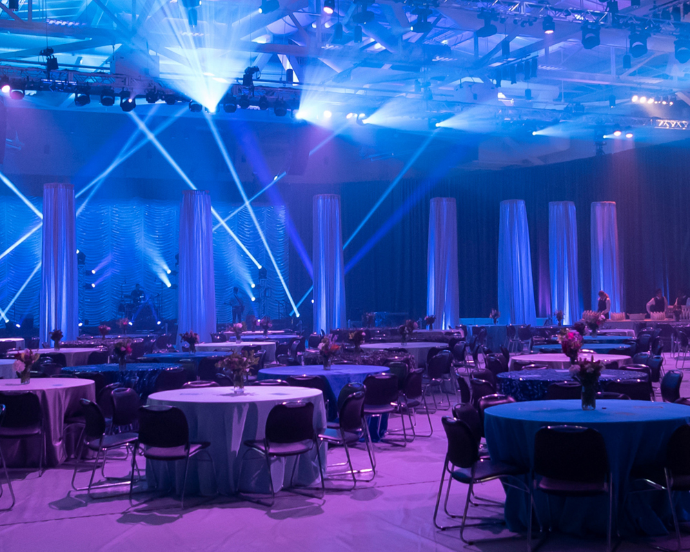 A large room being prepped for an event with round tables, centerpieces, and a band in the background