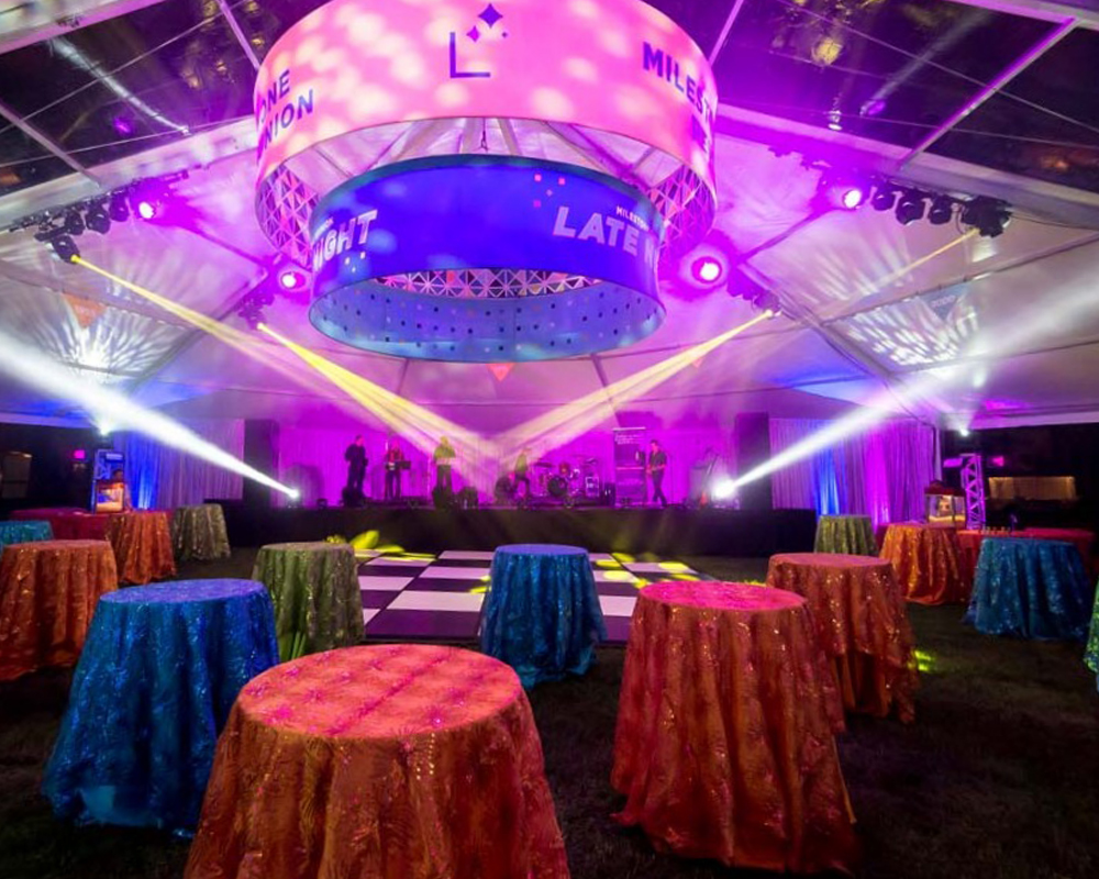 An outdoor event under a large tent, featuring colorful lights and decorations, and high top tables with colorful tablecloths
