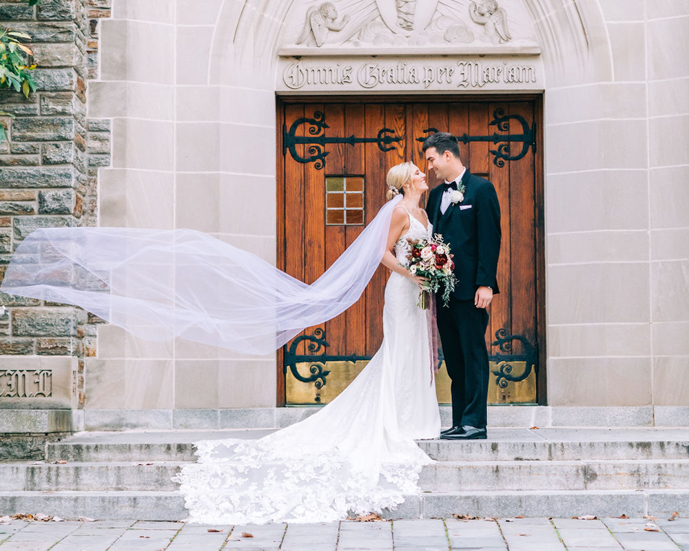 A couple posing for a photo in front of the Chapel doors with the wind elegantly blowing the brides train in the air