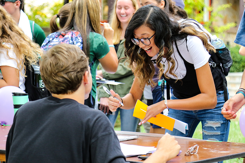 A smiling female student leans down to write on a table at the Activities Fair