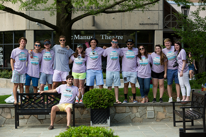 A group standing in a row in front of McManus Theatre, wearing matching shirts and with their arms across each other's shoulders