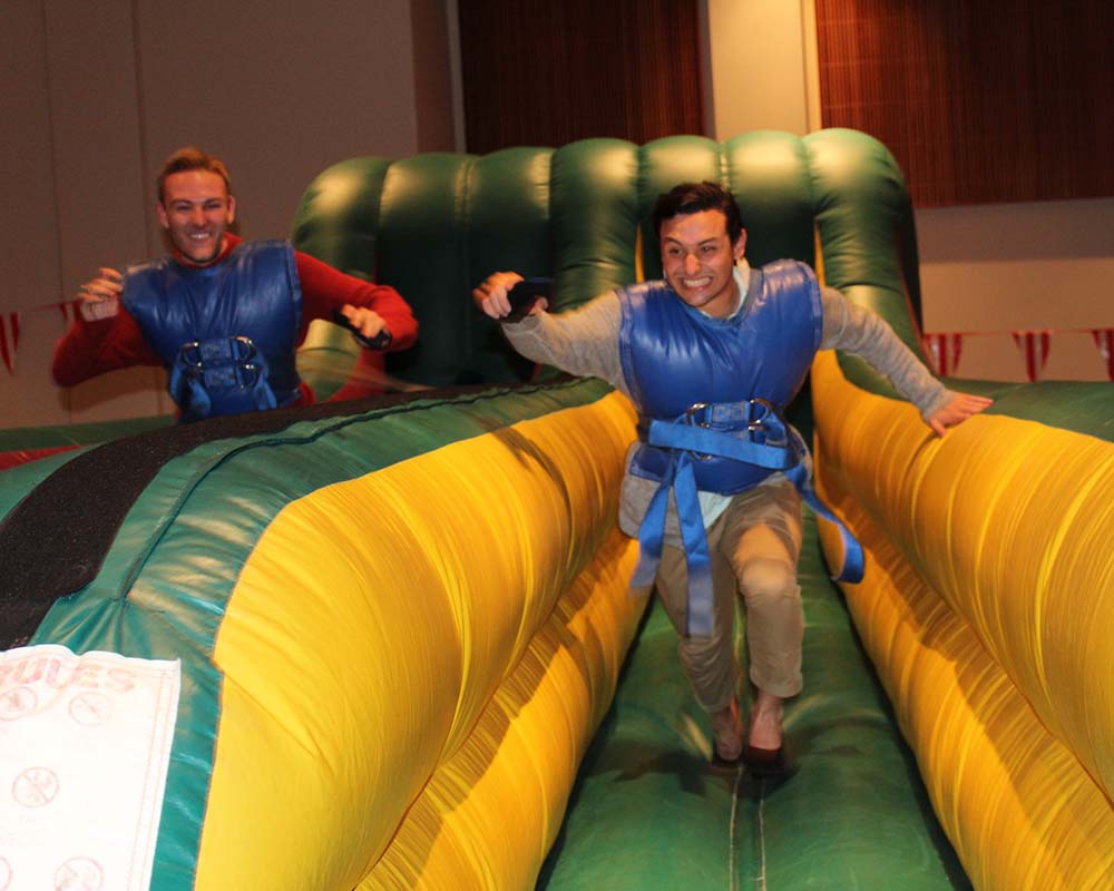 Two male students wearing padded vests run through an inflatable course