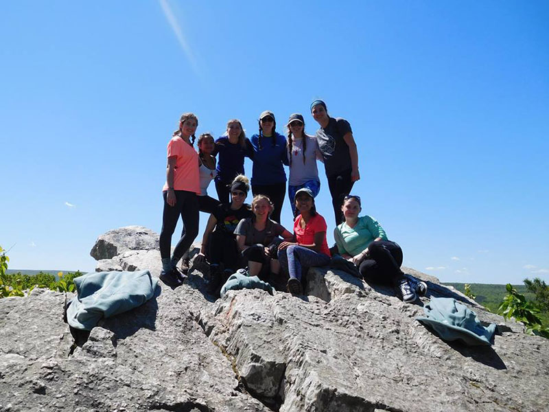 A group of female students posing for a photo on a large rock after a hike with clear blue skies