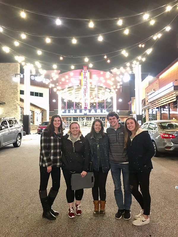 Students posing for a photo in the street in front of a movie theater, with lighting draped back and forth across the street from the building tops