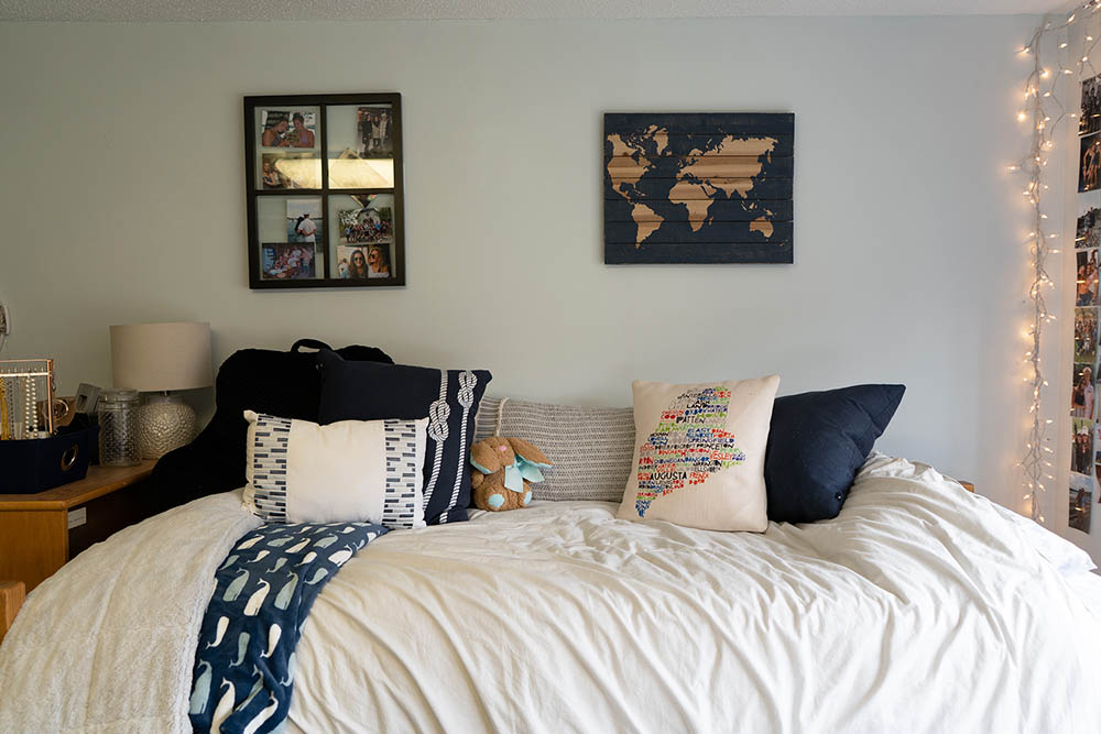 A dorm room bed with decorative pillows and frames on the wall behind it
