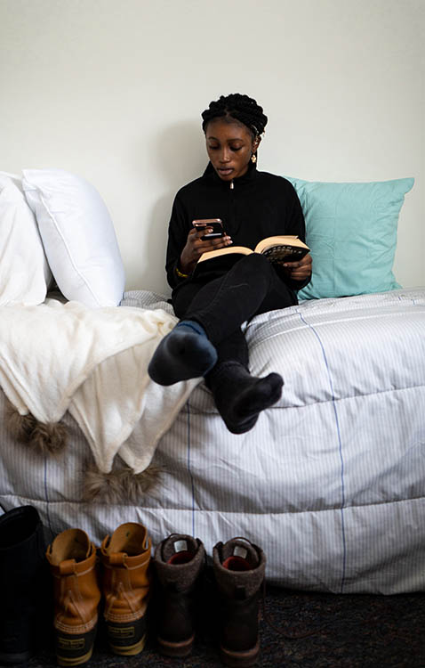 A student sits on their bed holding a book while reading her phone