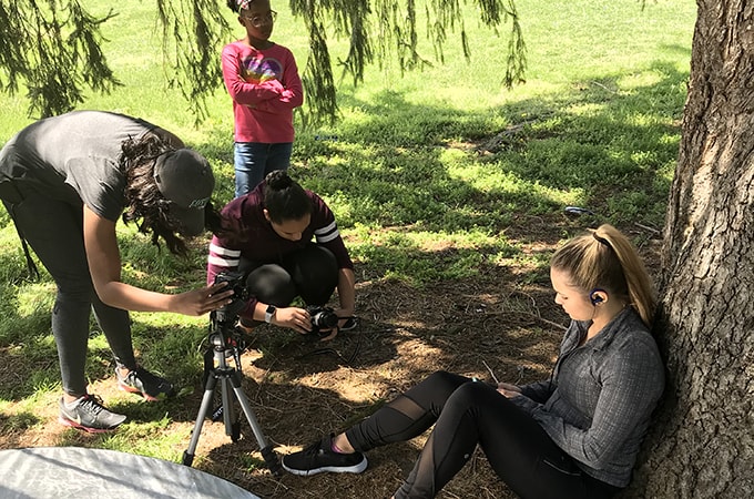 Two students filming another student sitting by a tree