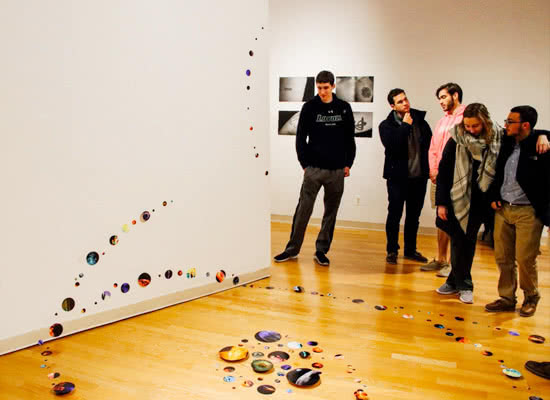 Visitors stand in an art gallery looking at artwork on the floor and walls