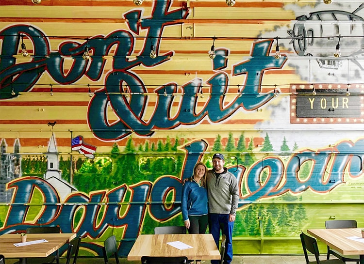 Loyola alumni Ryan and Alison Sampson Broderick pose in front of a painted mural that reads 'Don't Quit Your Daydream'
