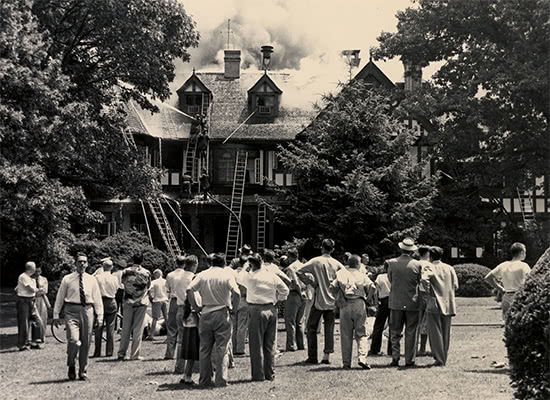Black and white photo of a large group of people watching firefighters extinguish a fire in the Humanities Building