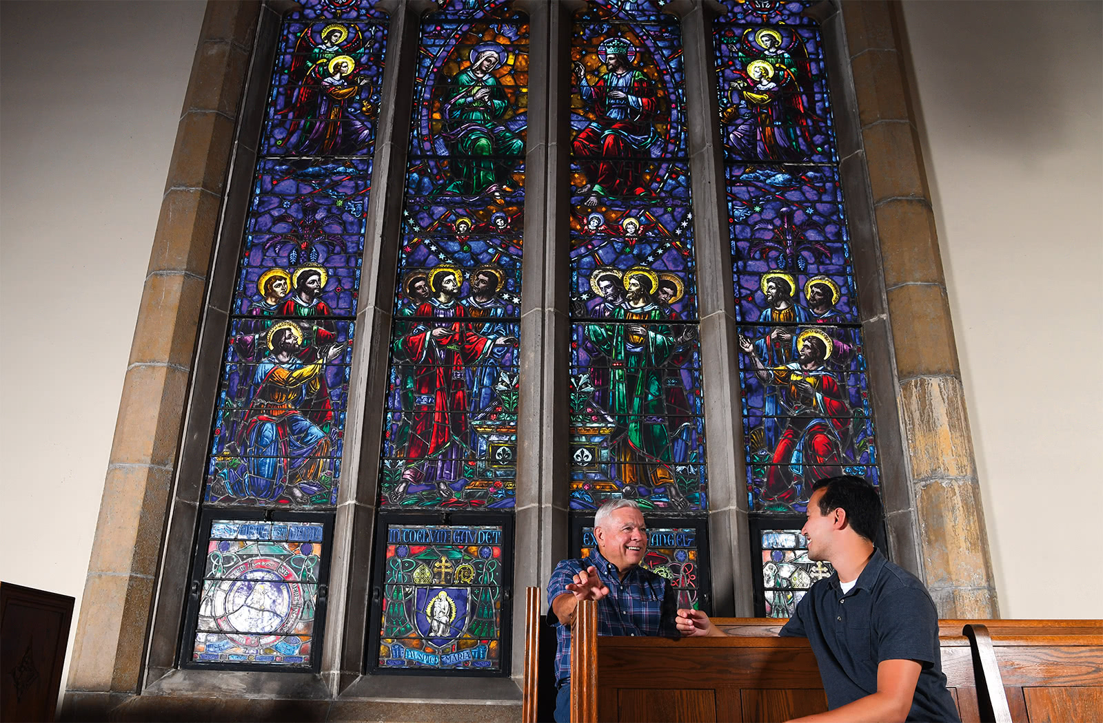 Two people sitting in front of a large, colorful, stained glass window talking and smiling