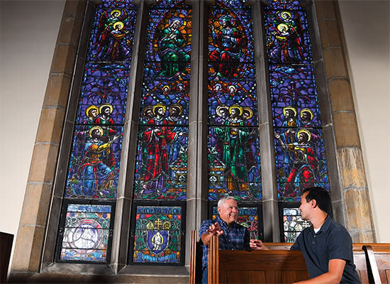 Two people sitting in front of a large, colorful, stained glass window talking and smiling
