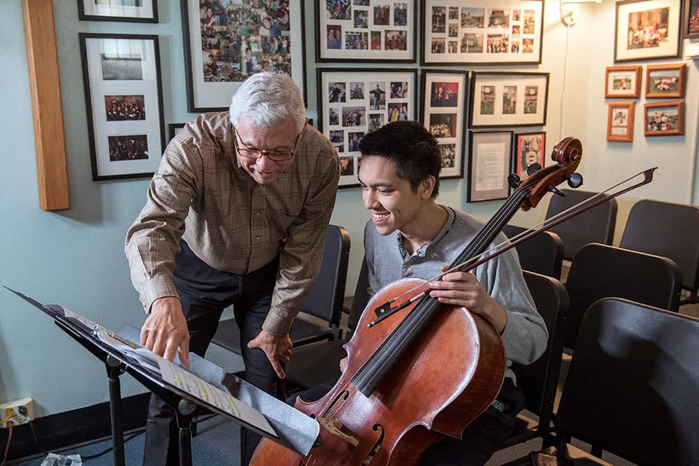 A professor points at music while a student sits nearby with a cello