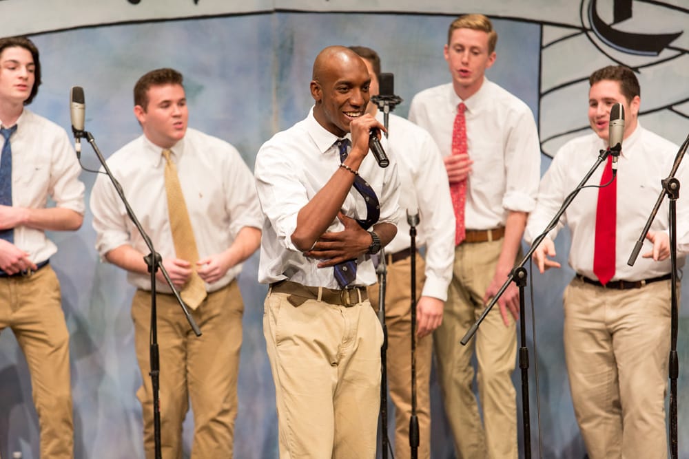A group of male students wearing tan pants and ties on stage singing, with one student in front singing a solo