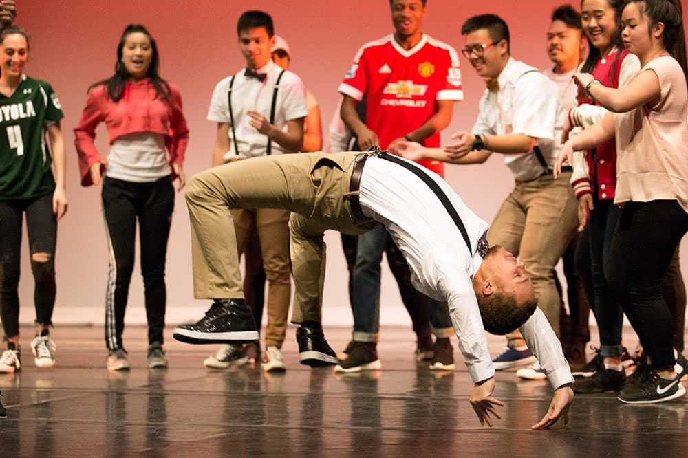 A student does a back flip on stage while other performing students cheer him on