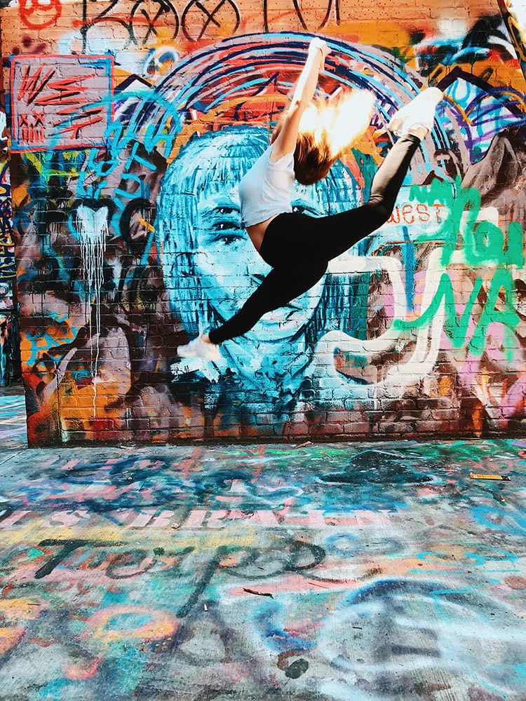 A student performs a dance jump in front of a wall with colorful graffiti