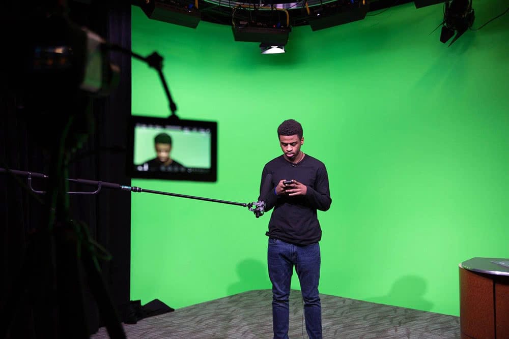 A student looking at a device while standing in front of a green screen