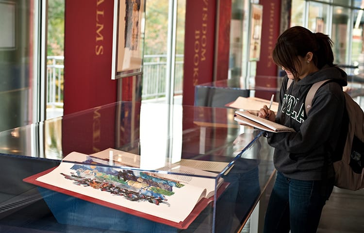 A female student taking notes in front of an exhibit with an illuminated manuscript