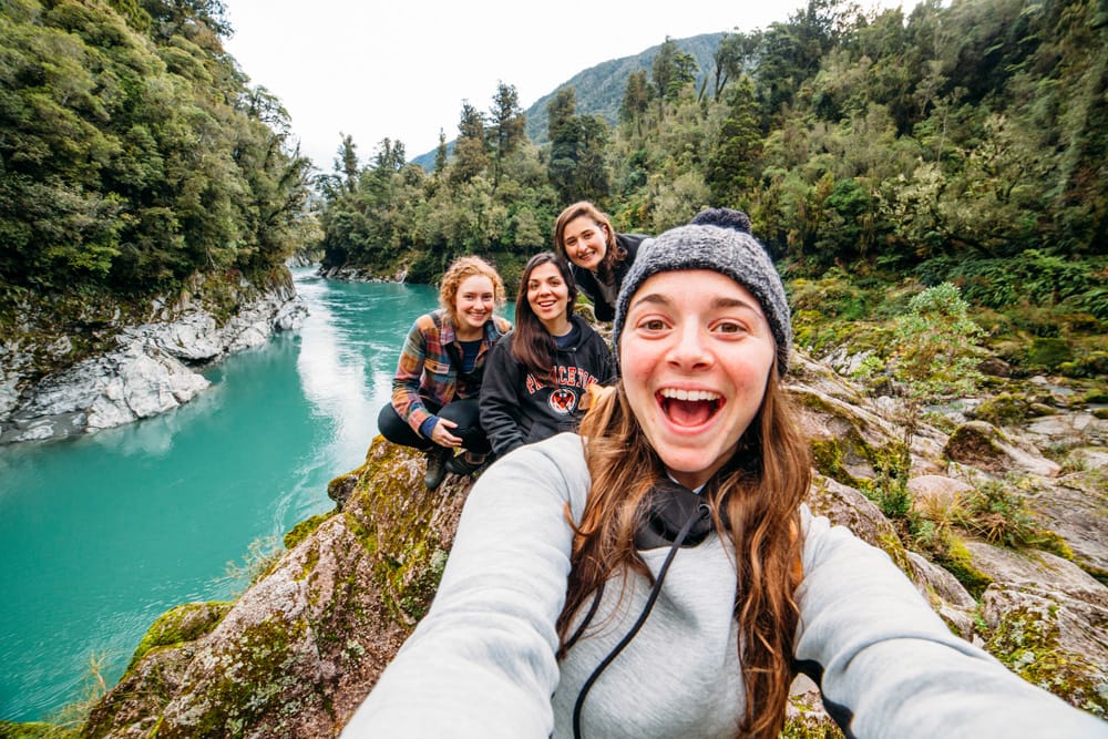 A student taking a selfie with other students in New Zealand - trees and a clear blue river can be seen in the background