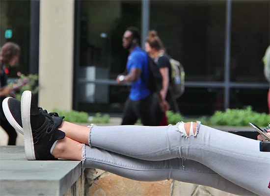 A students' legs propped up on an outdoor wall while studying and students pass by in the background