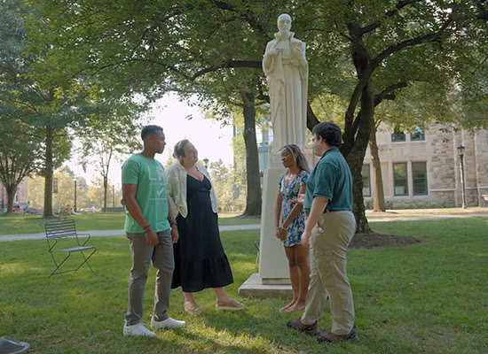 Students and staff chat while standing on Loyola's grassy academic quad, near a statue of St. Ignatius