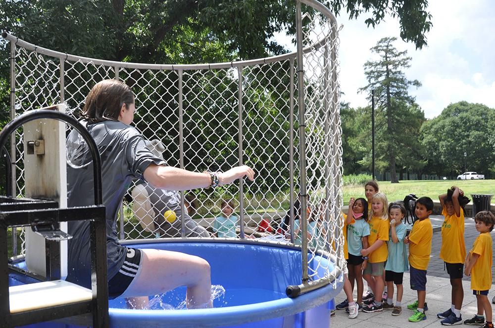 A drenched female counselor sits on a plank over a dunk tank while children line up for a chance to dunk her