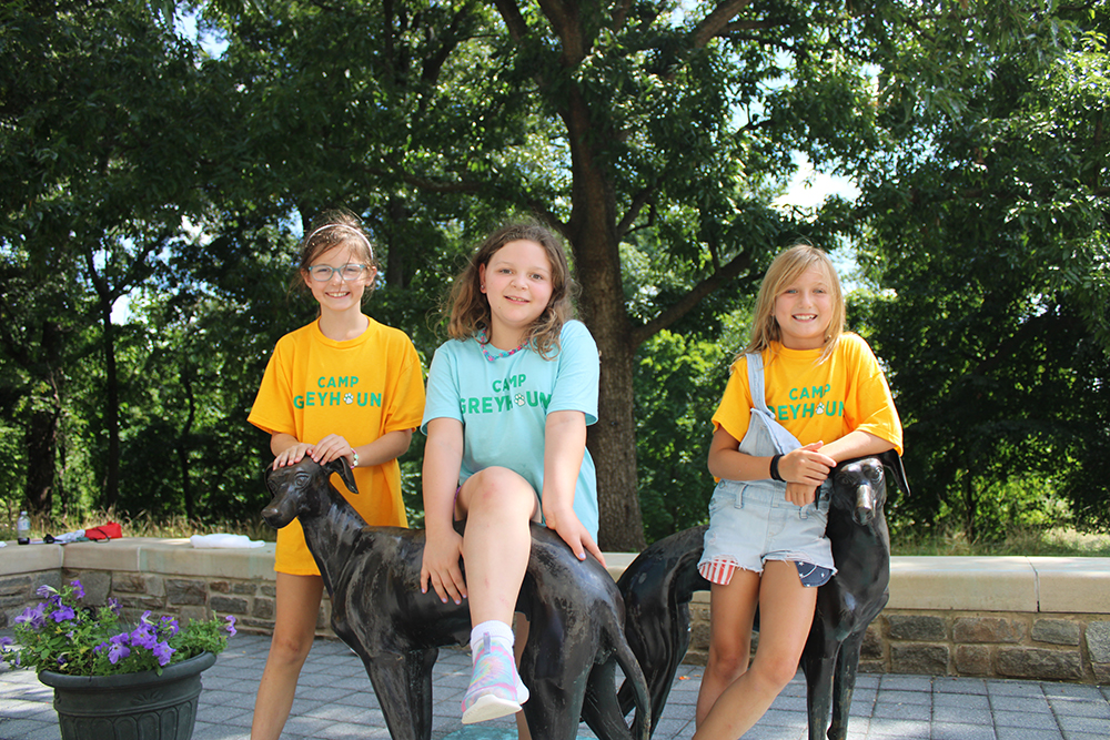 Three female campers pose for a photo with two Greyhound statues, Loyola's mascot