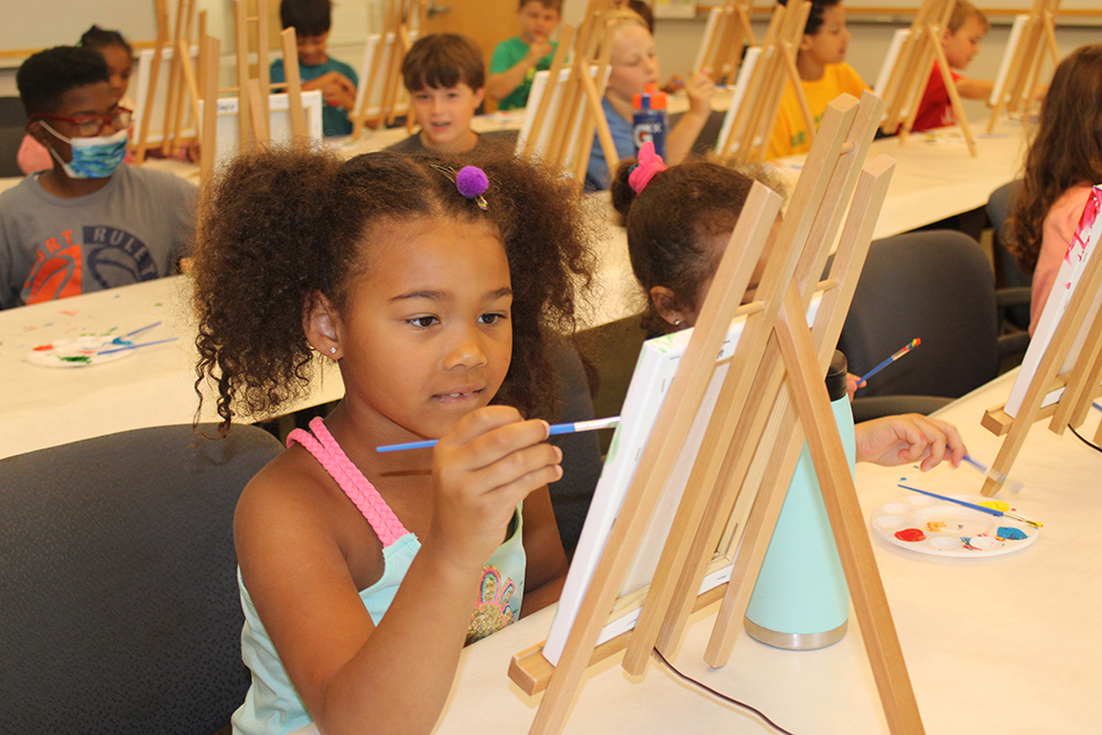 A female child paints a canvas on easel, with many more children doing the same in the background