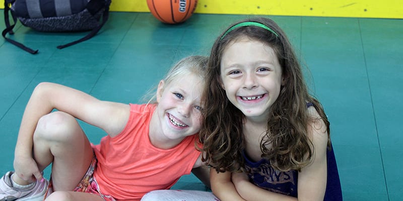Two female children leaning in for a picture together while sitting on a gym floor