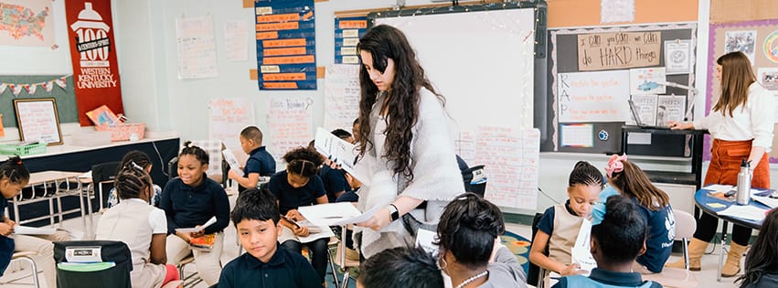 A female teacher hands out worksheets to a classroom of elementary students