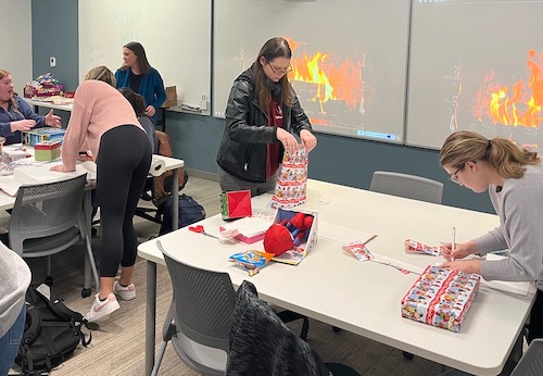 college students wrapping presents with a fireplace projected on a screen behind them