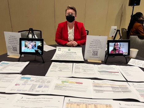 Woman wearing black KN95 mask and red blazer sitting at table with two iPads showing a picture of a mand and a woman. Papers are spread out on the table.