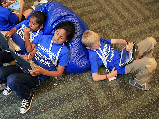 three students in the same blue t-shirts leaning against bean-bag chairs on computer tablets