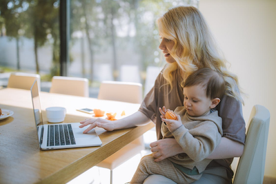 woman working on laptop with young child eating an orange on her lap
