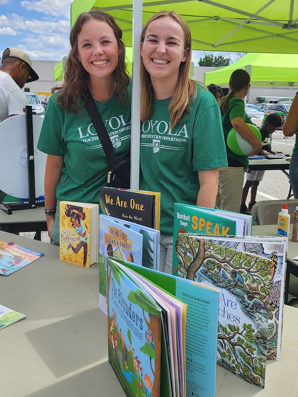 2 women with Loyola Teacher Education Department t-shirts stand in front of a table of books