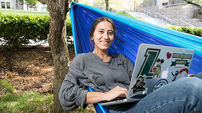 Student smiling at the camera as she uses her sticker-covered Macbook in a hammock on the quad