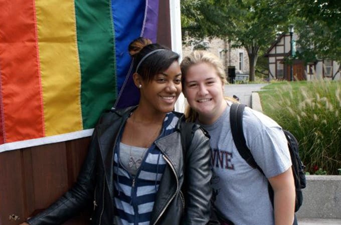 Two female students standing outside in a doorframe with a rainbow flag draped on the door