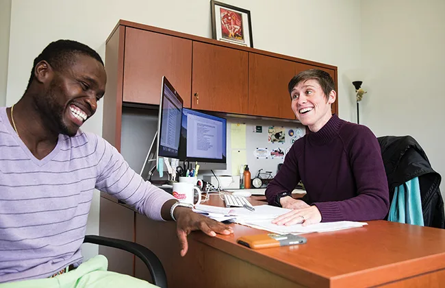 A student and professor laughing while sitting at a desk