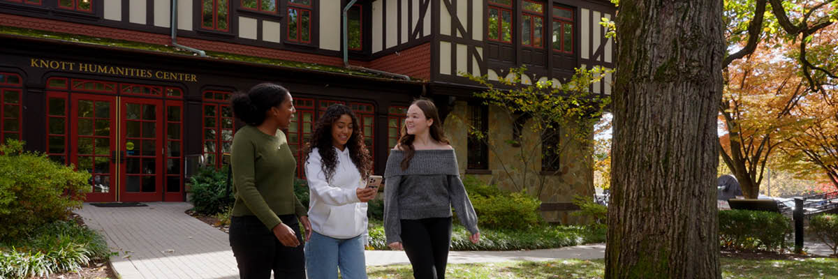 Three students walk and chat outside the Knott Humanities Center, surrounded by autumn trees.