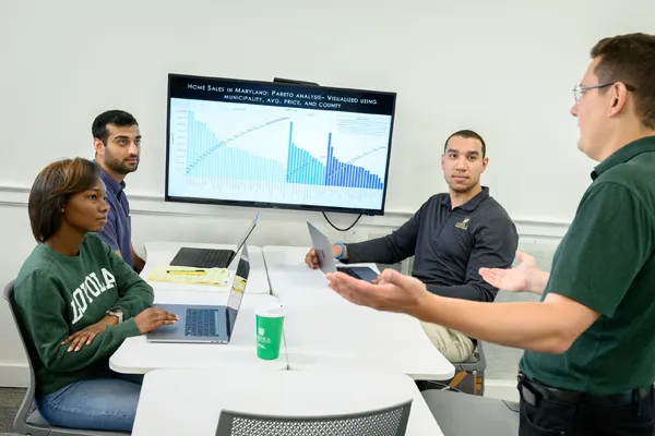 A professor talking with students sitting at a table in front of a monitor showing charts