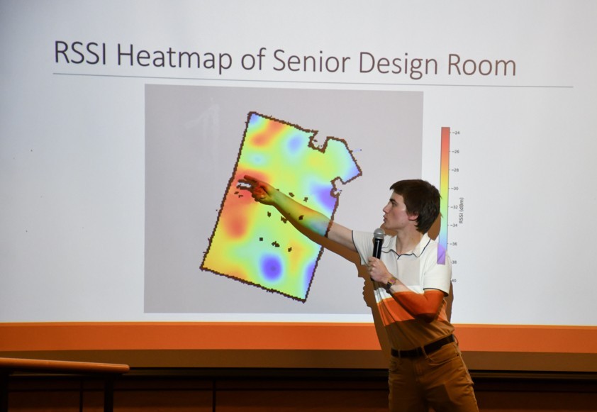 A student holds a microphone and points at a screen with a heat map graphic on it.