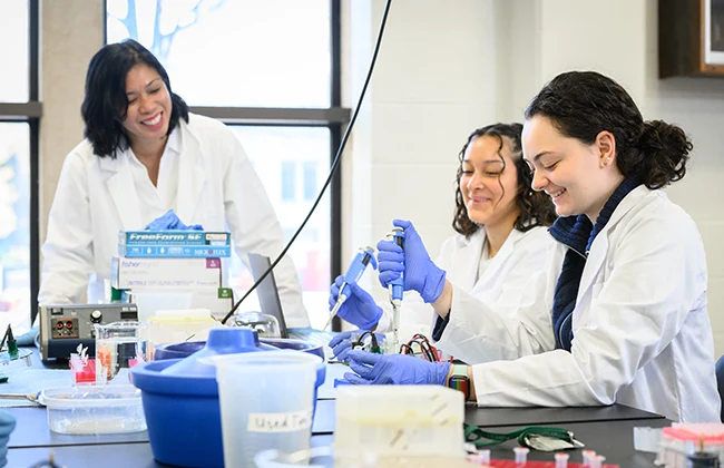 Two students in white lab coats work on an experiment while professor smiles and observes
