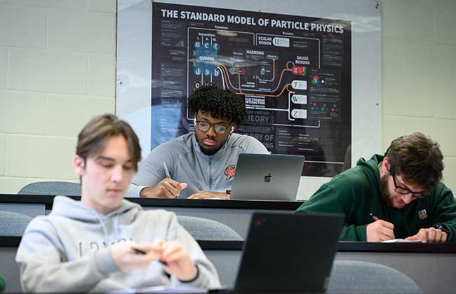 Students taking notes during a lecture with a particle physics poster on the wall