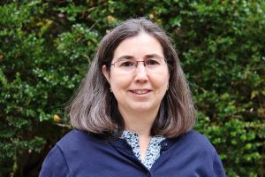 Ana Gomez-Perez from the shoulders up wearing a blue shirt in front of a green background